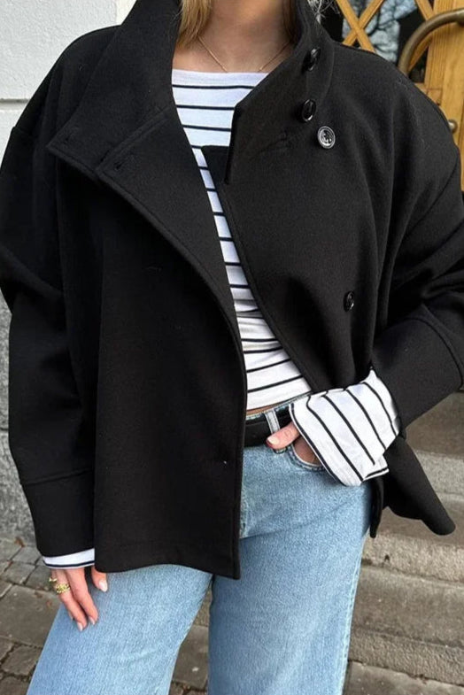 Person wearing a black coat over a striped shirt with light blue jeans on a street.